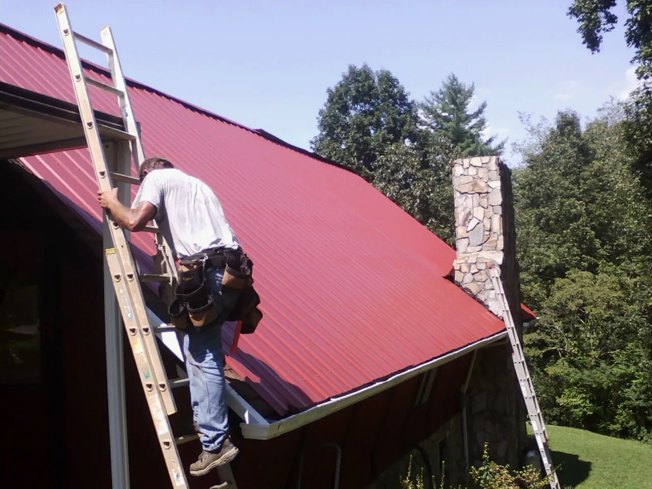 Roofer on a ladder beside a red metal roof and stone chimney — Soffit & Fascia Repair by Roofing Brothers in Teton